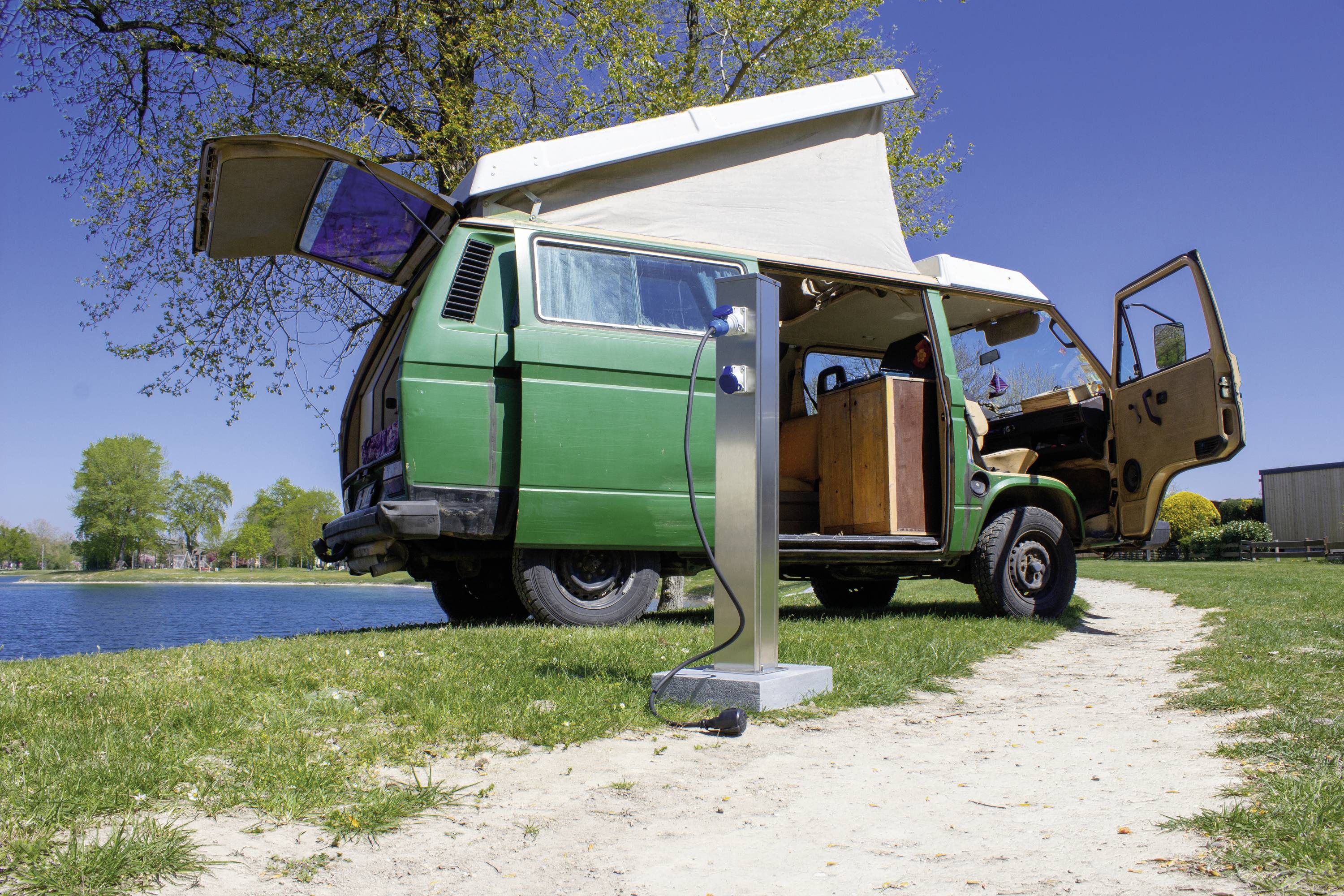 A green camper van with an open roof and doors is parked by the shore of a lake. One person sits inside the van, surrounded by trees.