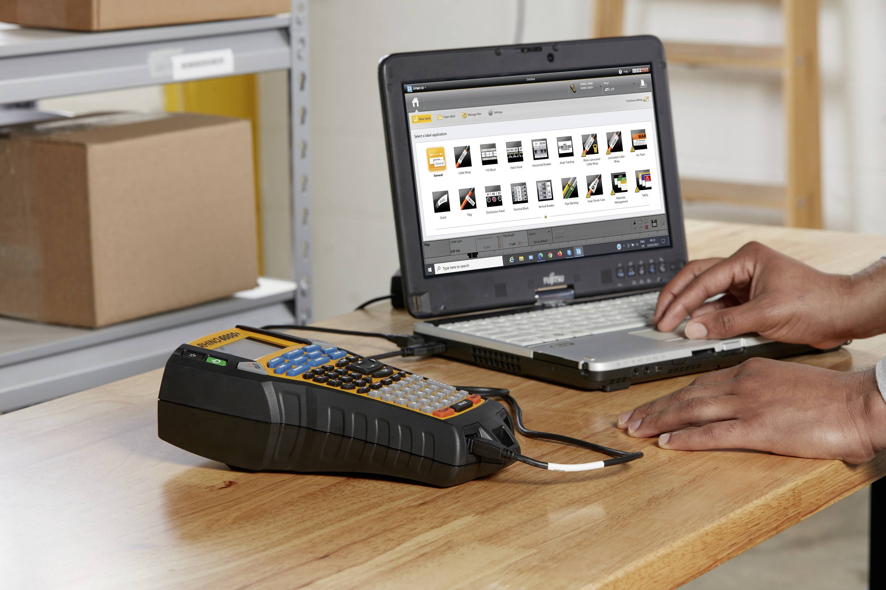 A person is working on a laptop at a wooden table with a label printer lying beside it. Shelves with cardboard boxes are in the background.