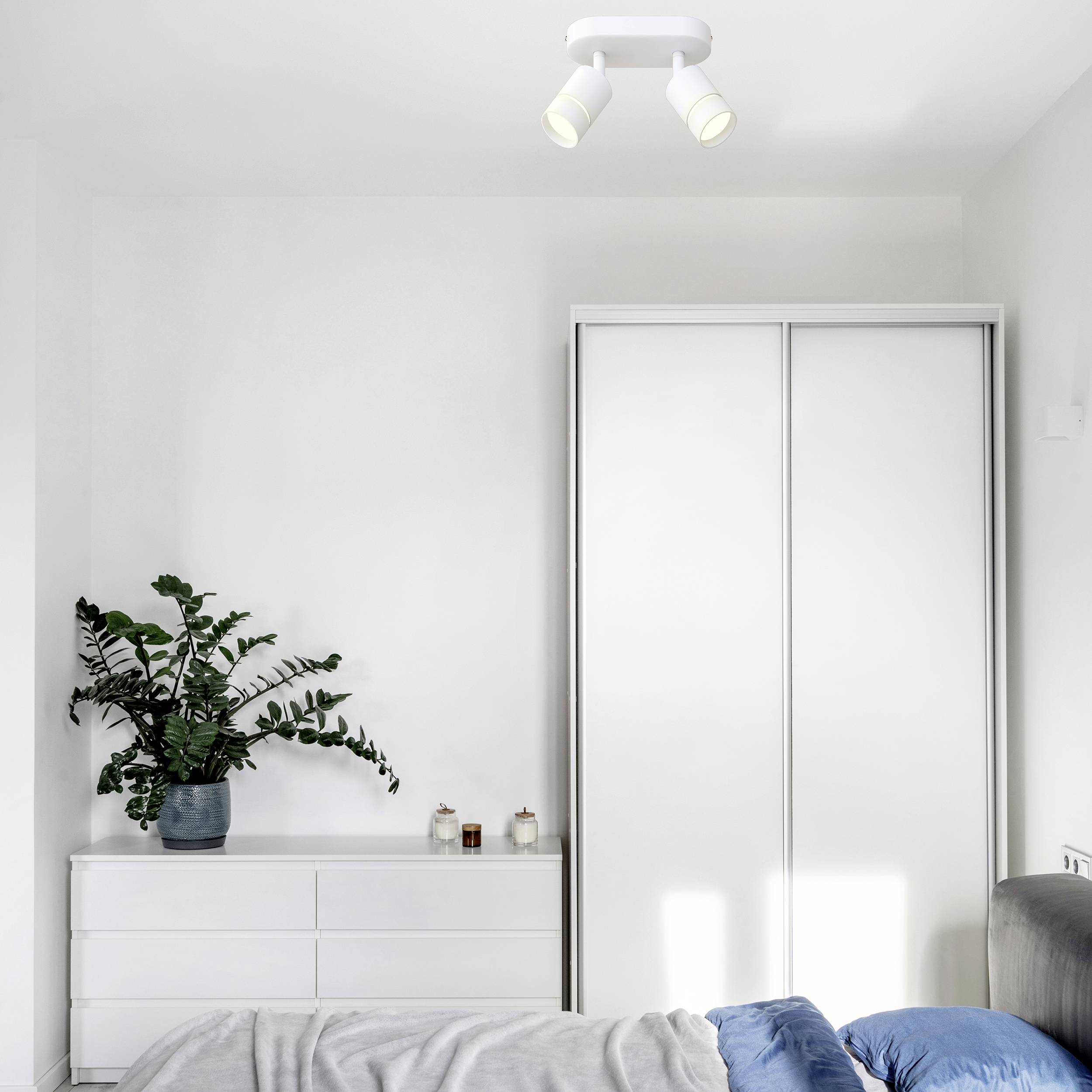A minimalist bedroom with a white wardrobe, a chest of drawers with plants, and a bed with a grey blanket and blue pillow.