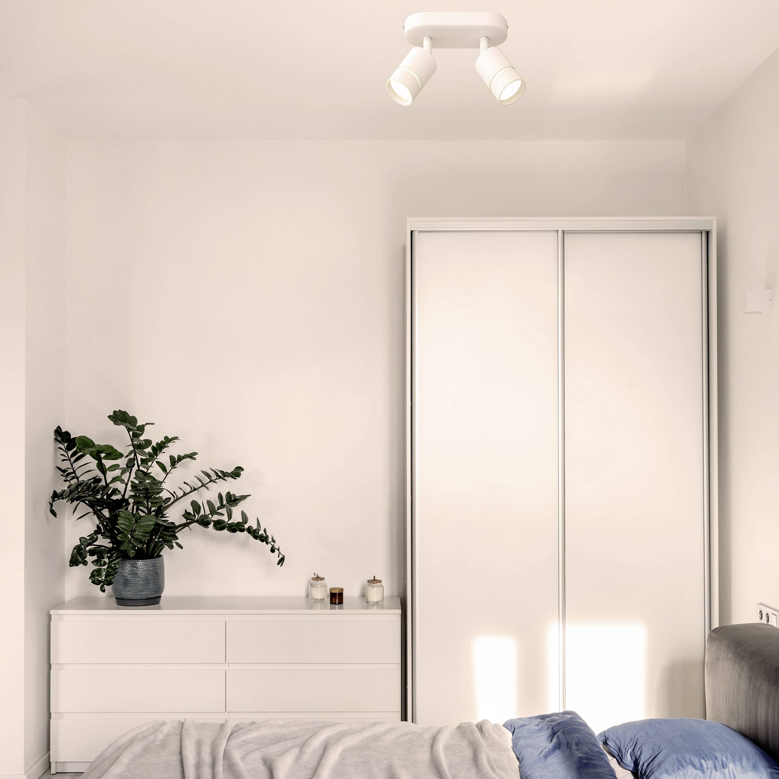 A simple bedroom with a white wardrobe, a chest of drawers, plants and a bed. Soft daylight illuminates the room.