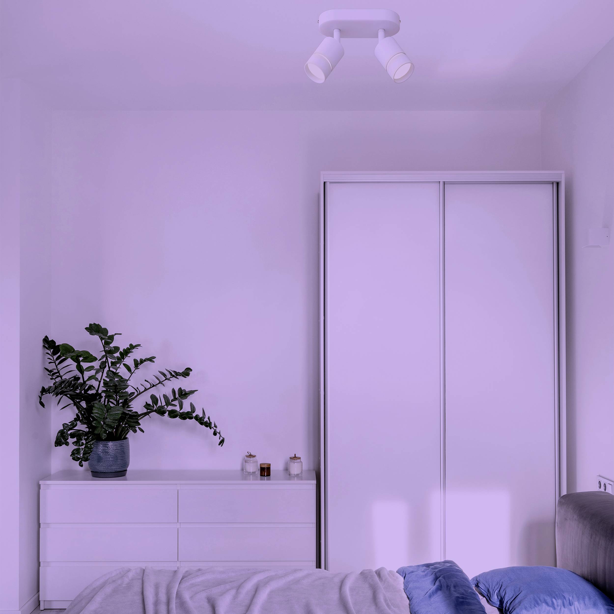 A bedroom with a white wardrobe, a chest of drawers with a plant and small bottles. A bed with a grey blanket in the foreground.