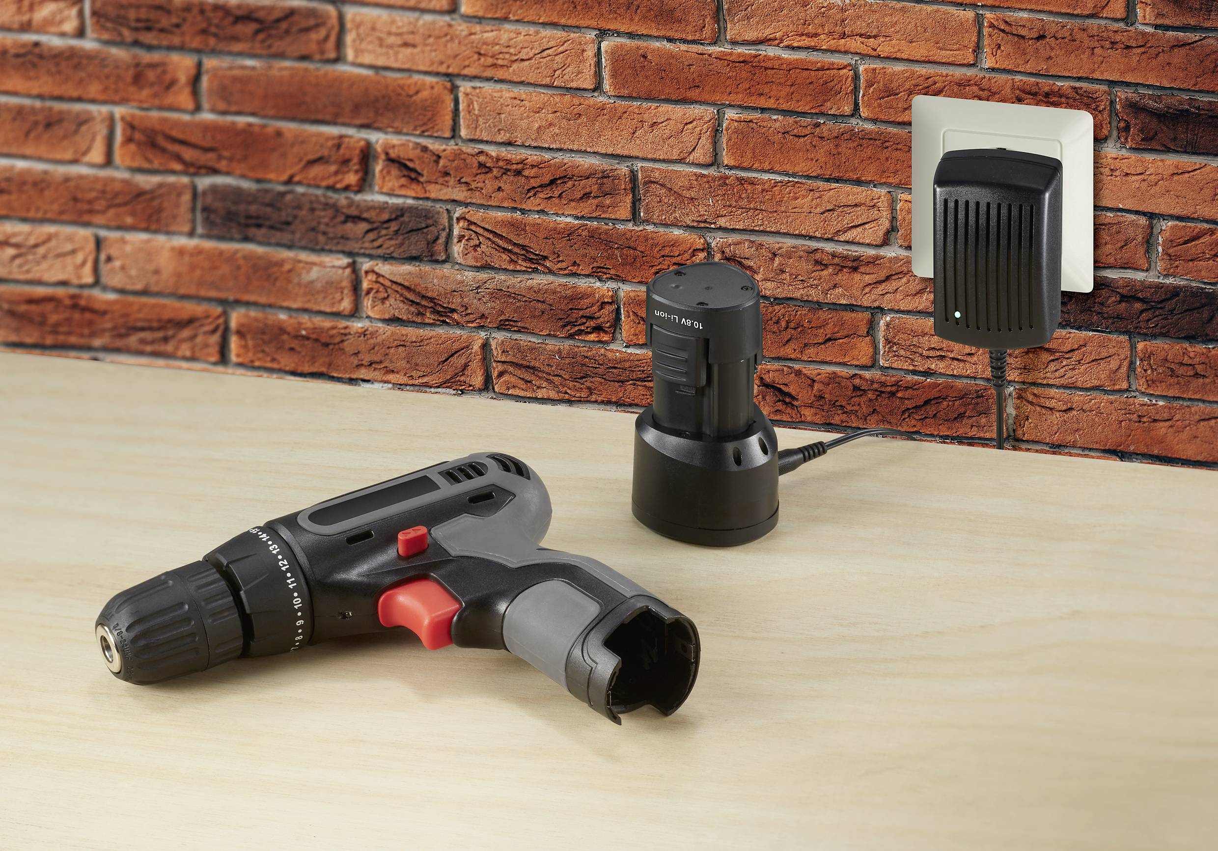 Cordless drill lies on a table next to its charger, which is plugged into a socket on a brick wall.