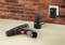 Cordless drill lies on a table next to its charger, which is plugged into a socket on a brick wall.