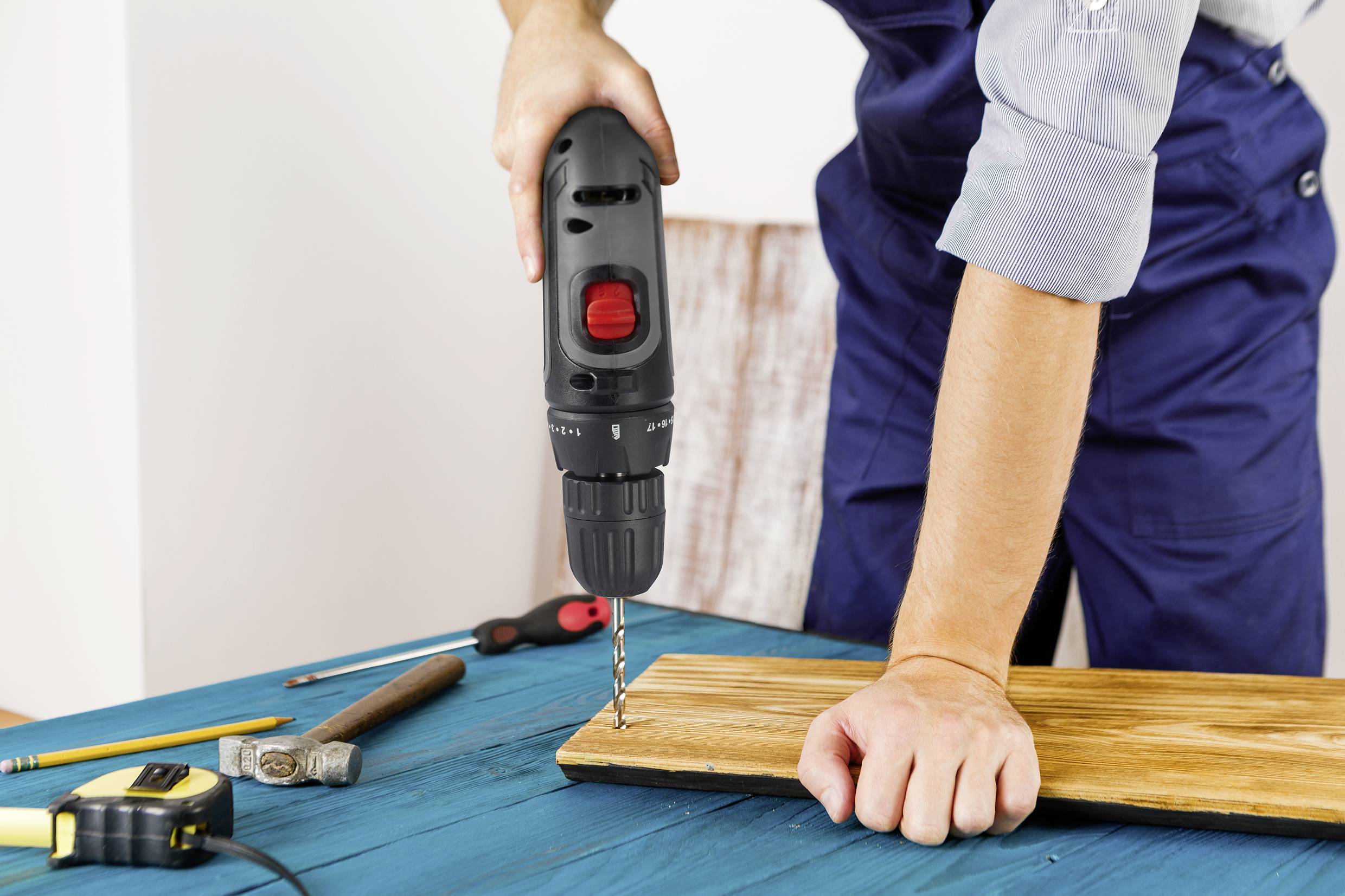 A person is drilling into a wooden board with a cordless drill, which is lying on a table. Tools are placed beside it.