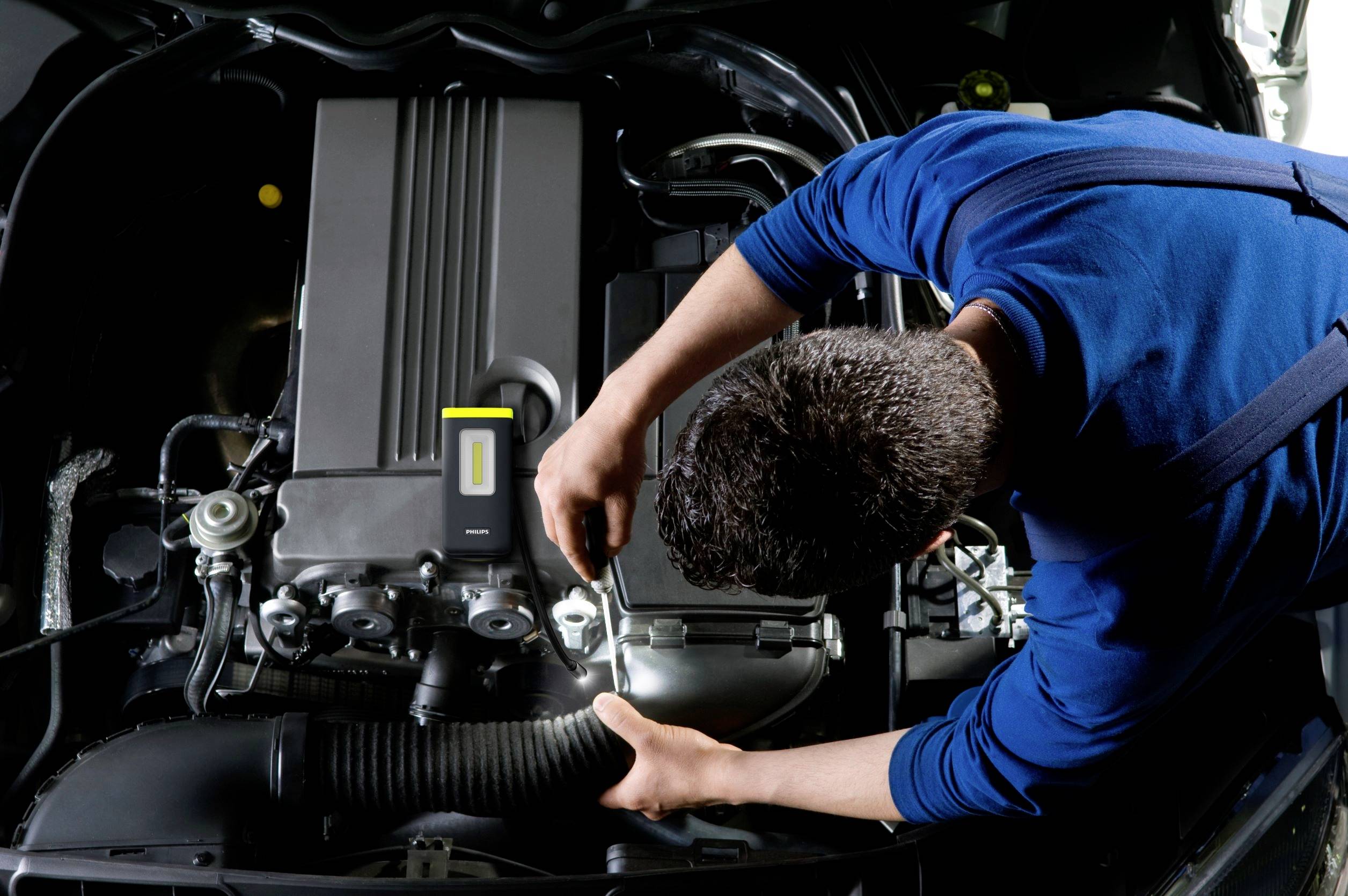 A mechanic is working under the raised bonnet of a car and tightening a screw. The engine is clearly visible.