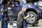 A mechanic is checking the tyre of a raised blue car in a workshop.