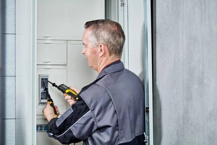 An electrician in work attire is working with a screwdriver on an open electrical distribution box inside a building.