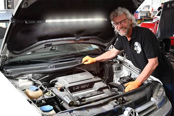 A person is smiling while working on an open car engine in a workshop. They are wearing orange gloves and a black T-shirt.