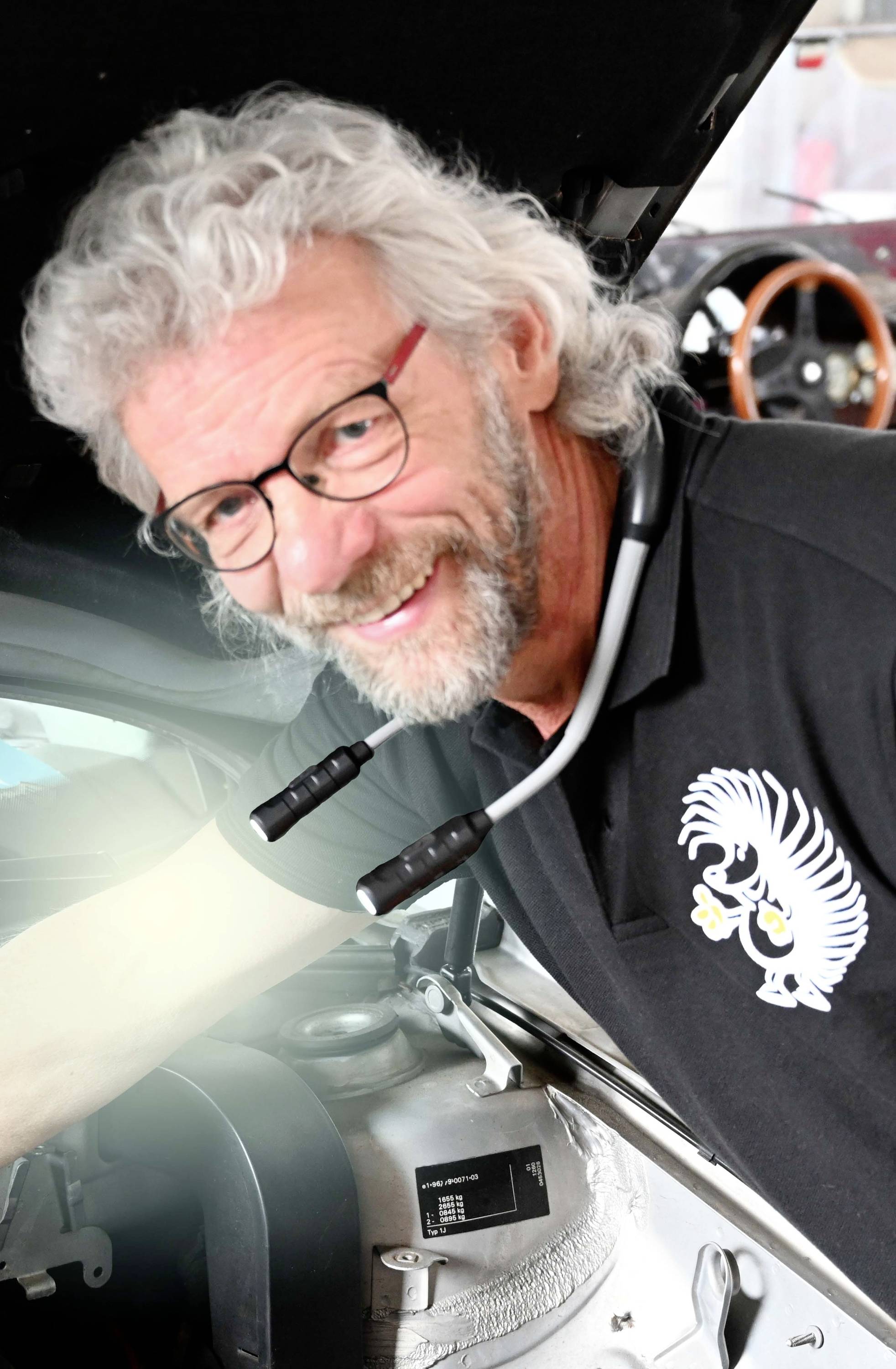 A person with grey hair and glasses smiles while looking under the raised bonnet of a car.