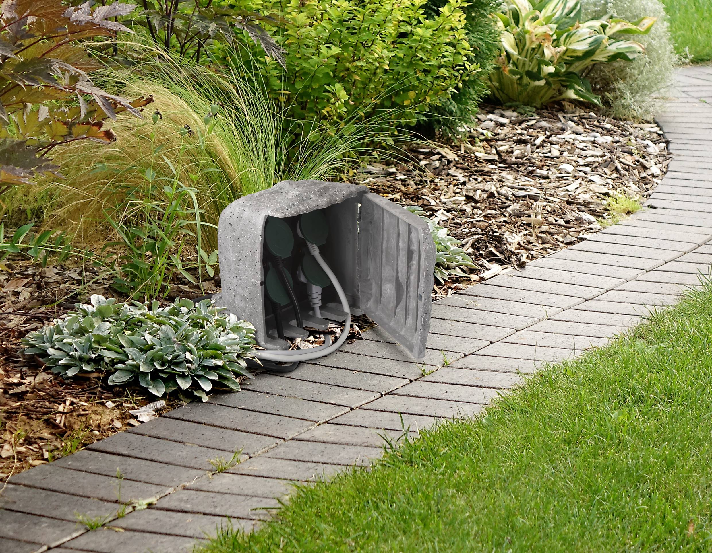 A grey electrical socket with an open junction box in the garden, surrounded by plants and a paved pathway.