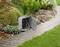 A grey electrical socket with an open junction box in the garden, surrounded by plants and a paved pathway.