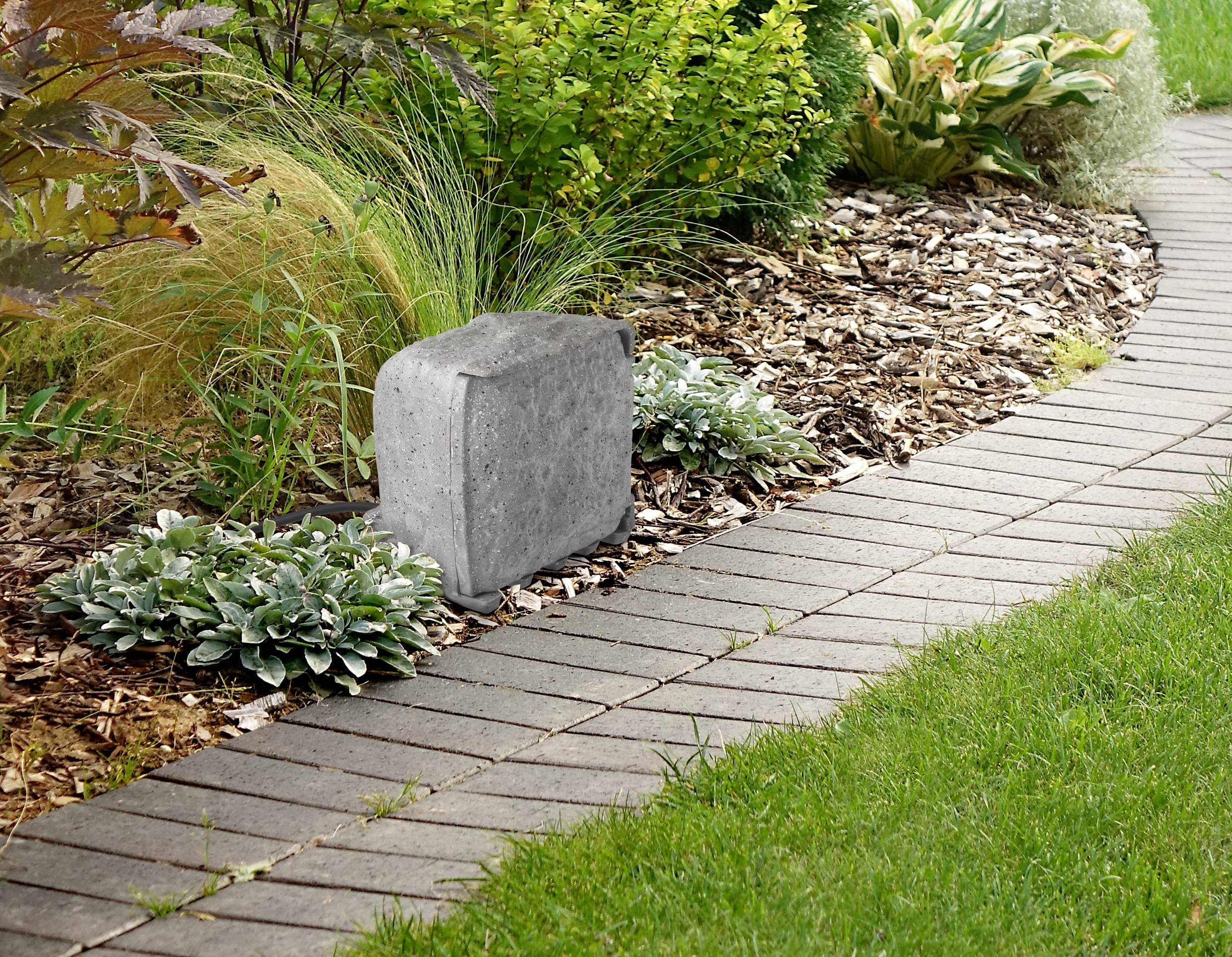A grey stone lies in a well-maintained bed with ground-covering plants and grasses beside a paved path.
