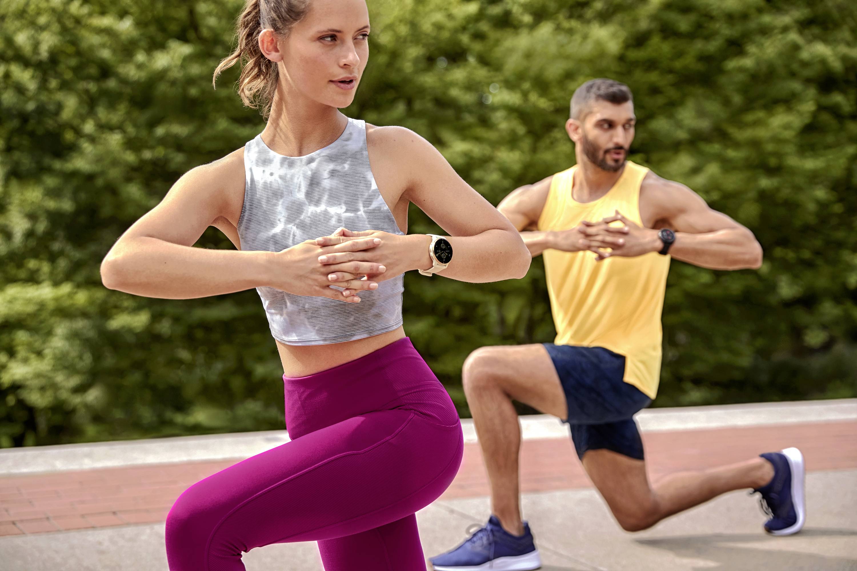 Two people in sportswear are doing lunges in a park. The woman is wearing a grey top and pink leggings, the man a yellow shirt.