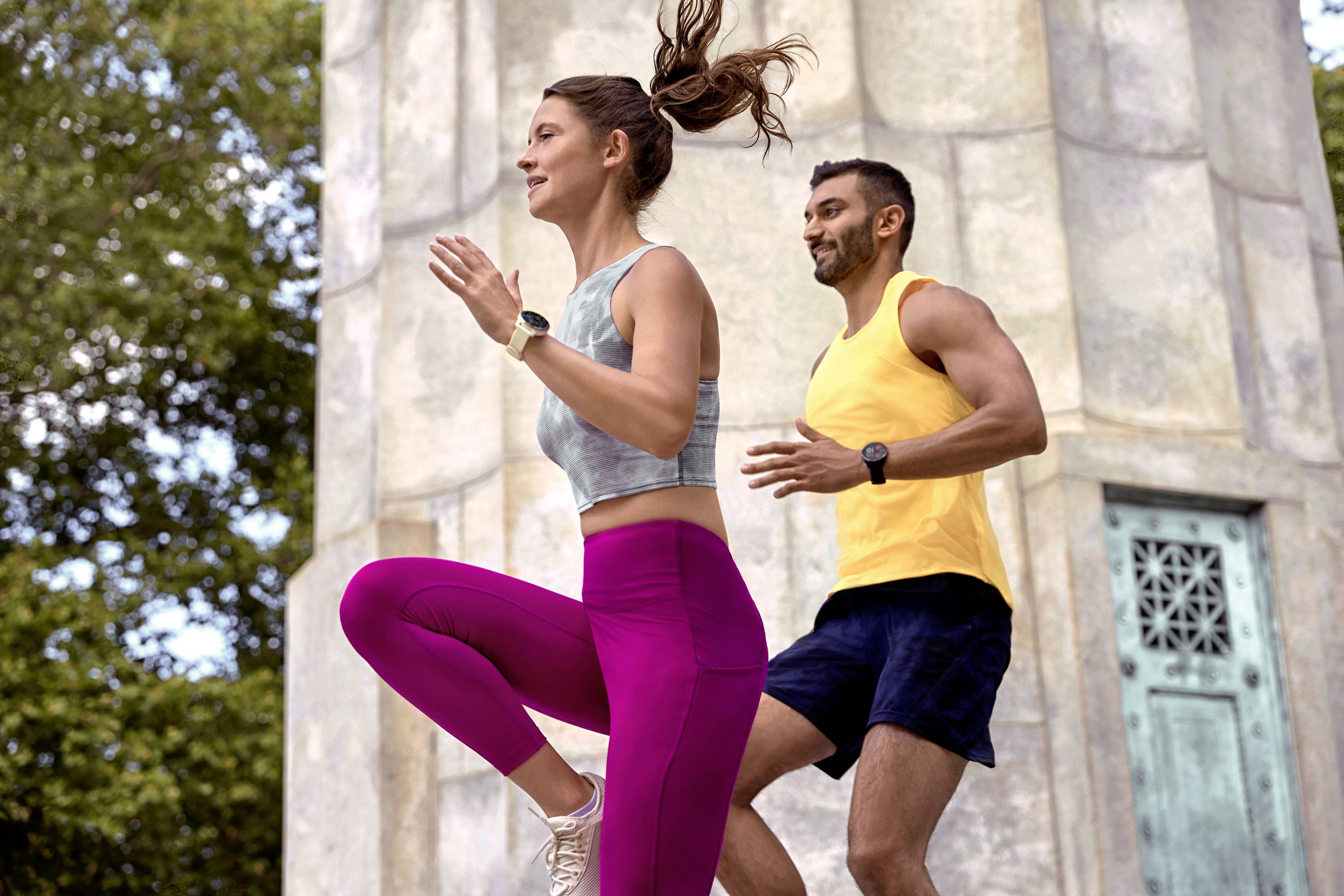 A woman and a man are doing fitness exercises outside next to a stone column. They are wearing sports clothing and appear focused.