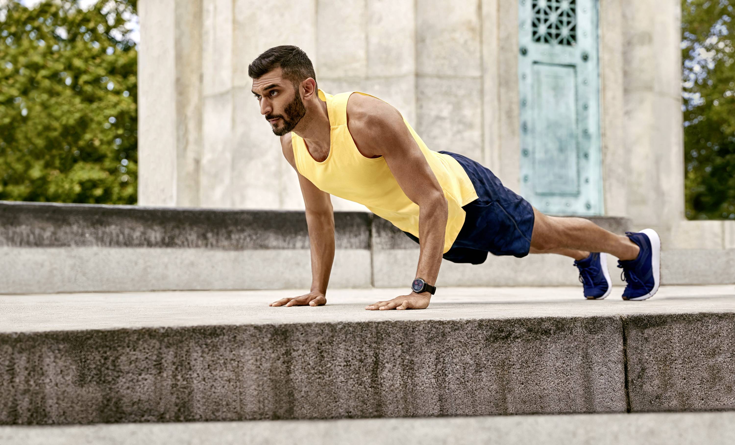 A man in a yellow vest and blue shorts is doing press-ups on an outdoor stone platform.