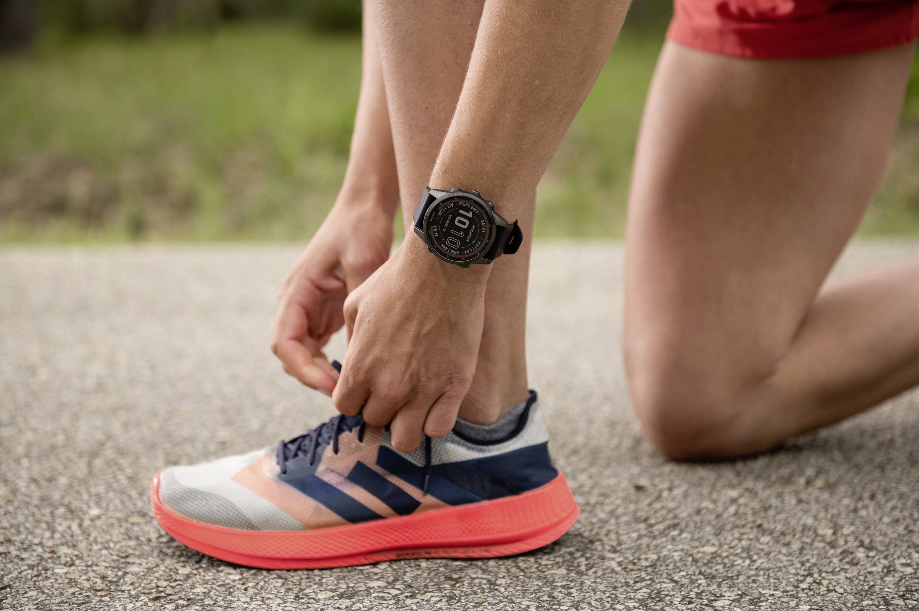 A person is kneeling on a path and tying the laces of a trainer. The person is wearing a watch and is dressed in sportswear.