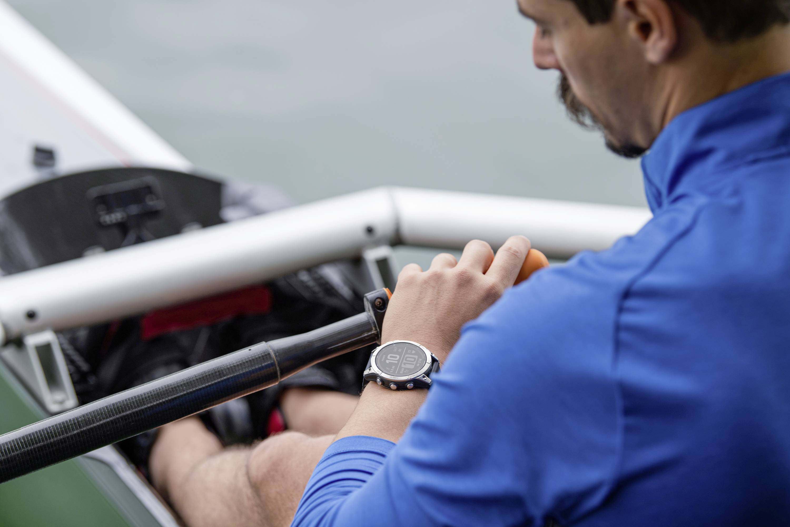 A man in a blue jacket is rowing in a boat, wearing a smartwatch on his wrist. Focus on the watch and the rowing motion.