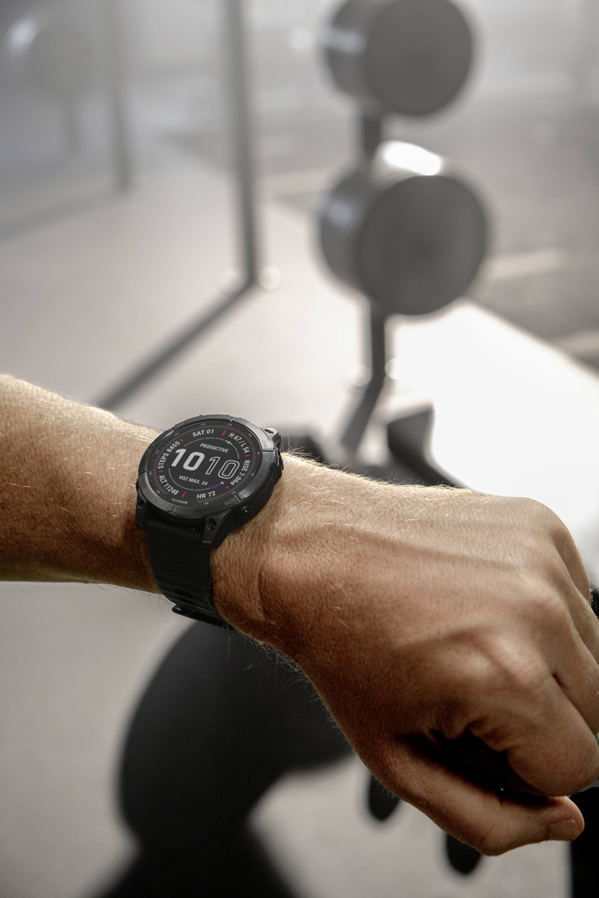 A person is wearing a smartwatch showing 10:10, standing in a gym in front of a blurred dumbbell rack.