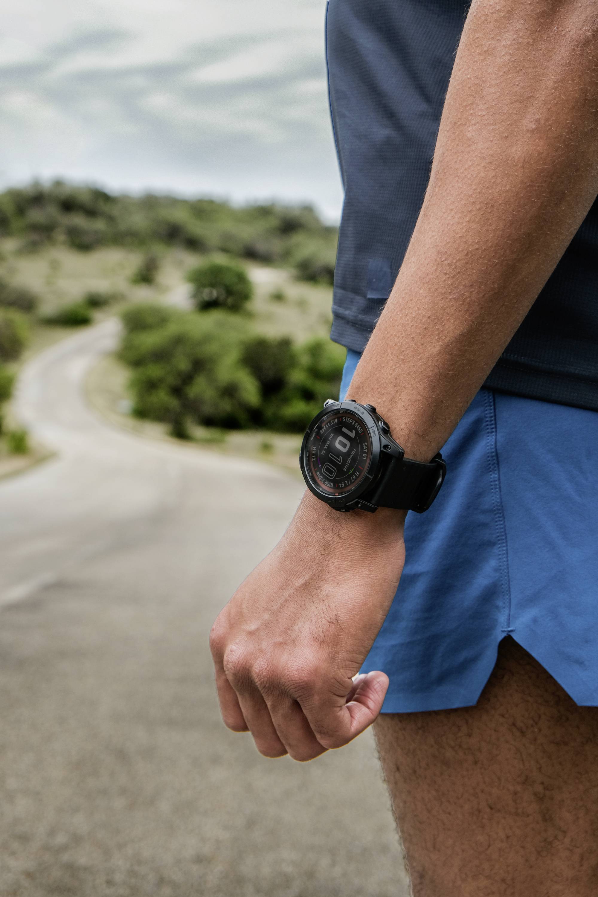 A person is jogging along a rural road, wearing blue shorts and a dark grey shirt, with a large black wristwatch on their wrist.