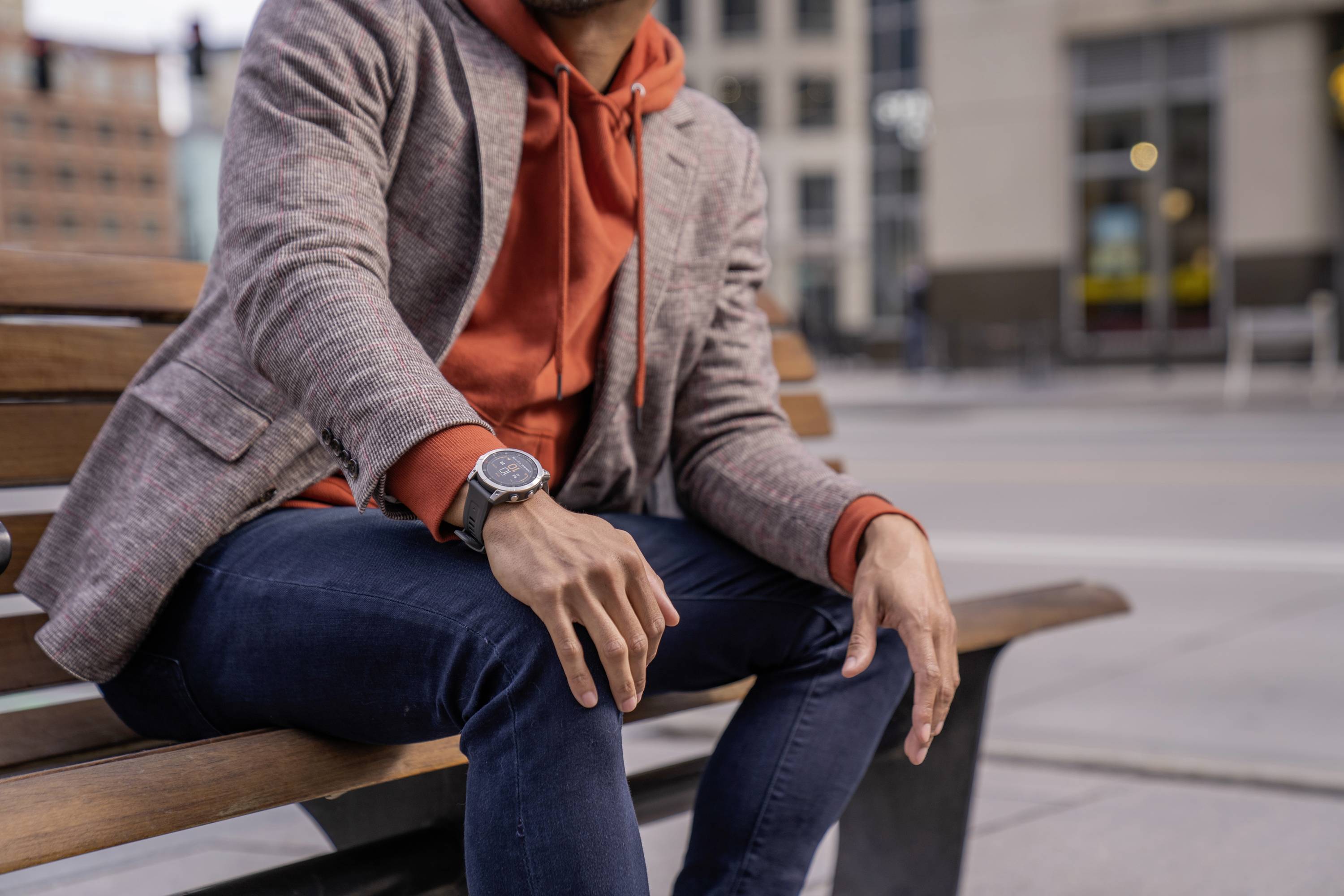 A person is sitting relaxed on a bench in the city, wearing an orange hoodie under a grey blazer and a wristwatch.