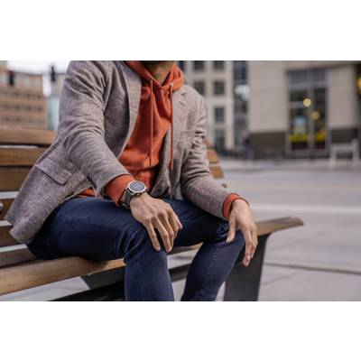 A person is sitting relaxed on a bench in the city, wearing an orange hoodie under a grey blazer and a wristwatch.