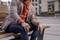 A person is sitting relaxed on a bench in the city, wearing an orange hoodie under a grey blazer and a wristwatch.