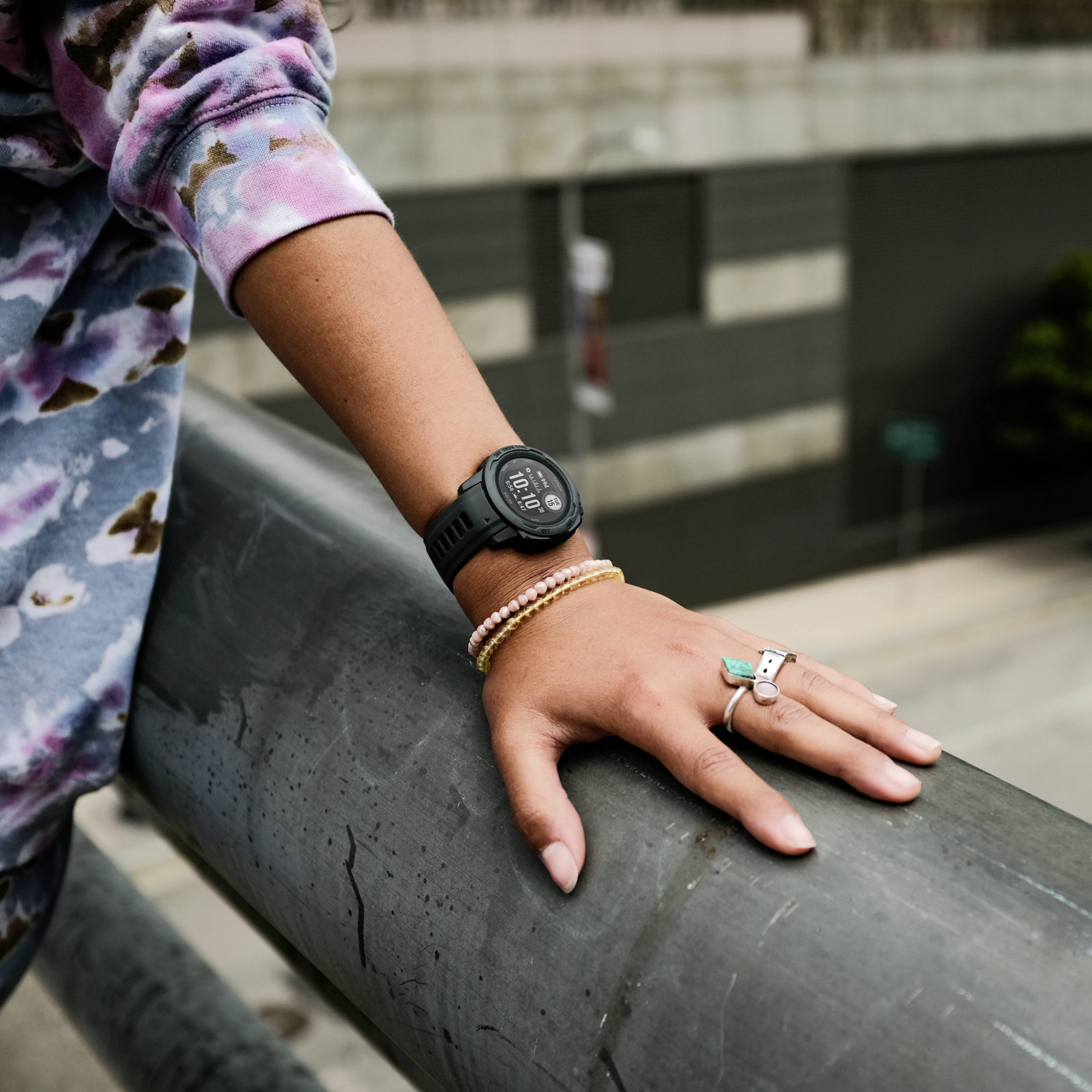 A person wearing colourful clothing leans against a railing. They are wearing a black watch on their wrist and have jewellery on their hand.