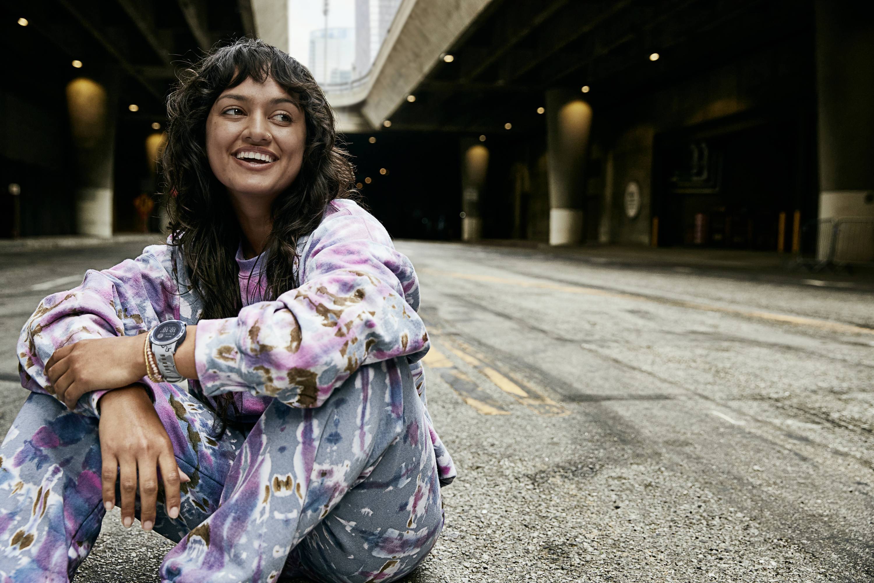 A smiling person sits relaxed on a street beneath a bridge. They are wearing colourful clothing and looking to the side.