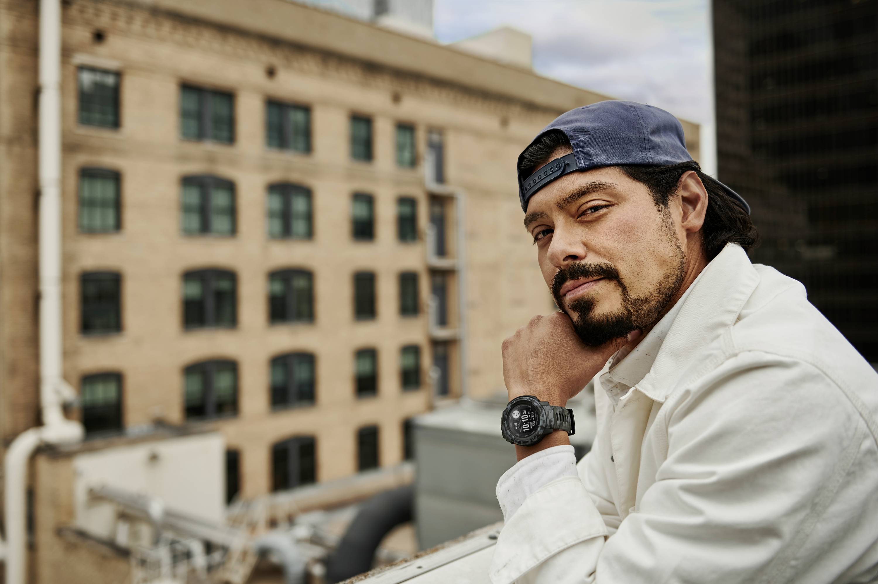 A man in a white jacket and blue cap is leaning on a railing in an urban setting with brick buildings in the background.