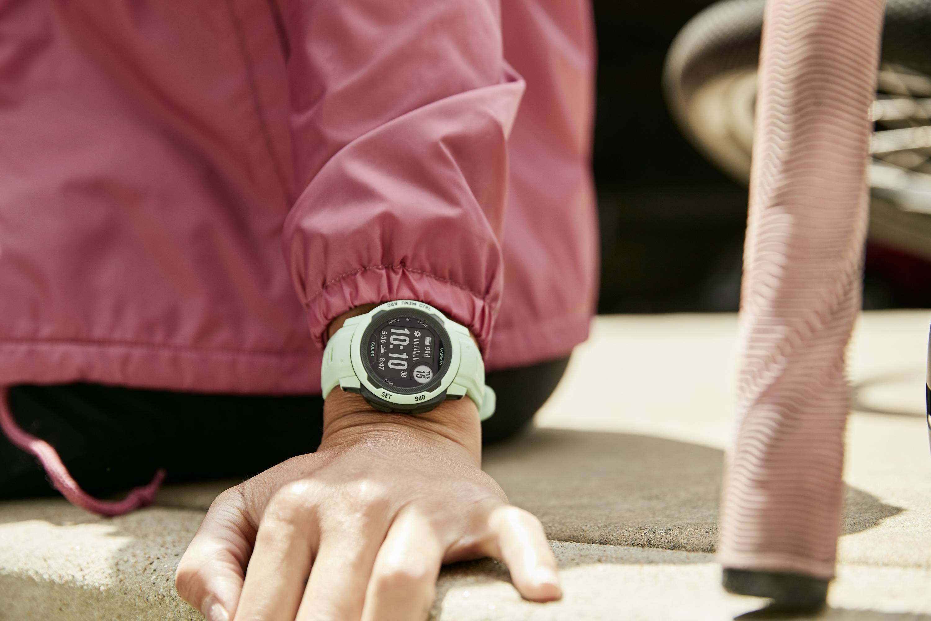 A person sitting on a step, wearing a smartwatch on their wrist. The watch displays the time and fitness data. A bicycle handlebar is visible in the background.