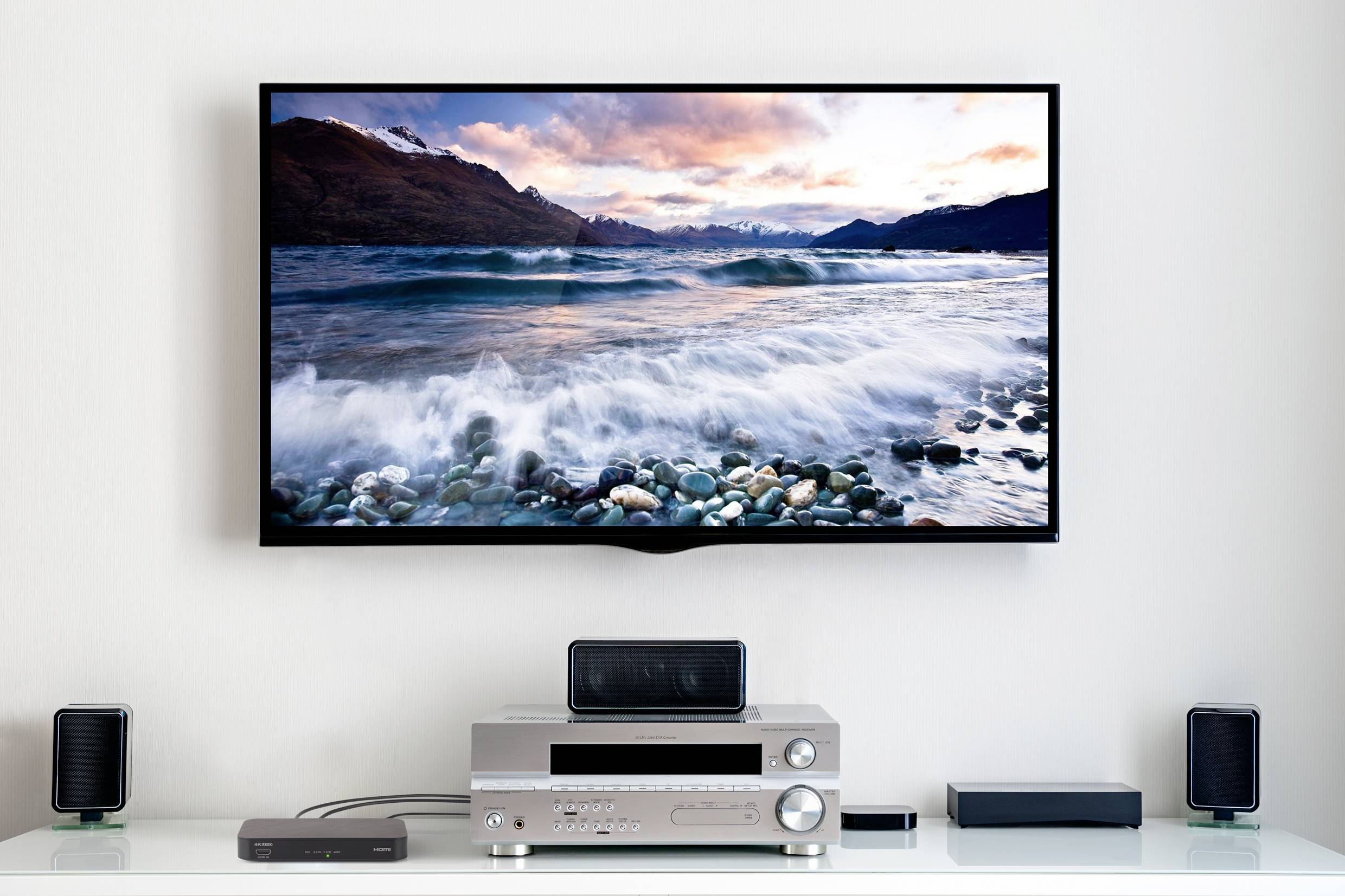 A home cinema setup with a flatscreen television mounted on the wall, displaying a landscape with water and mountains, surrounded by speakers.