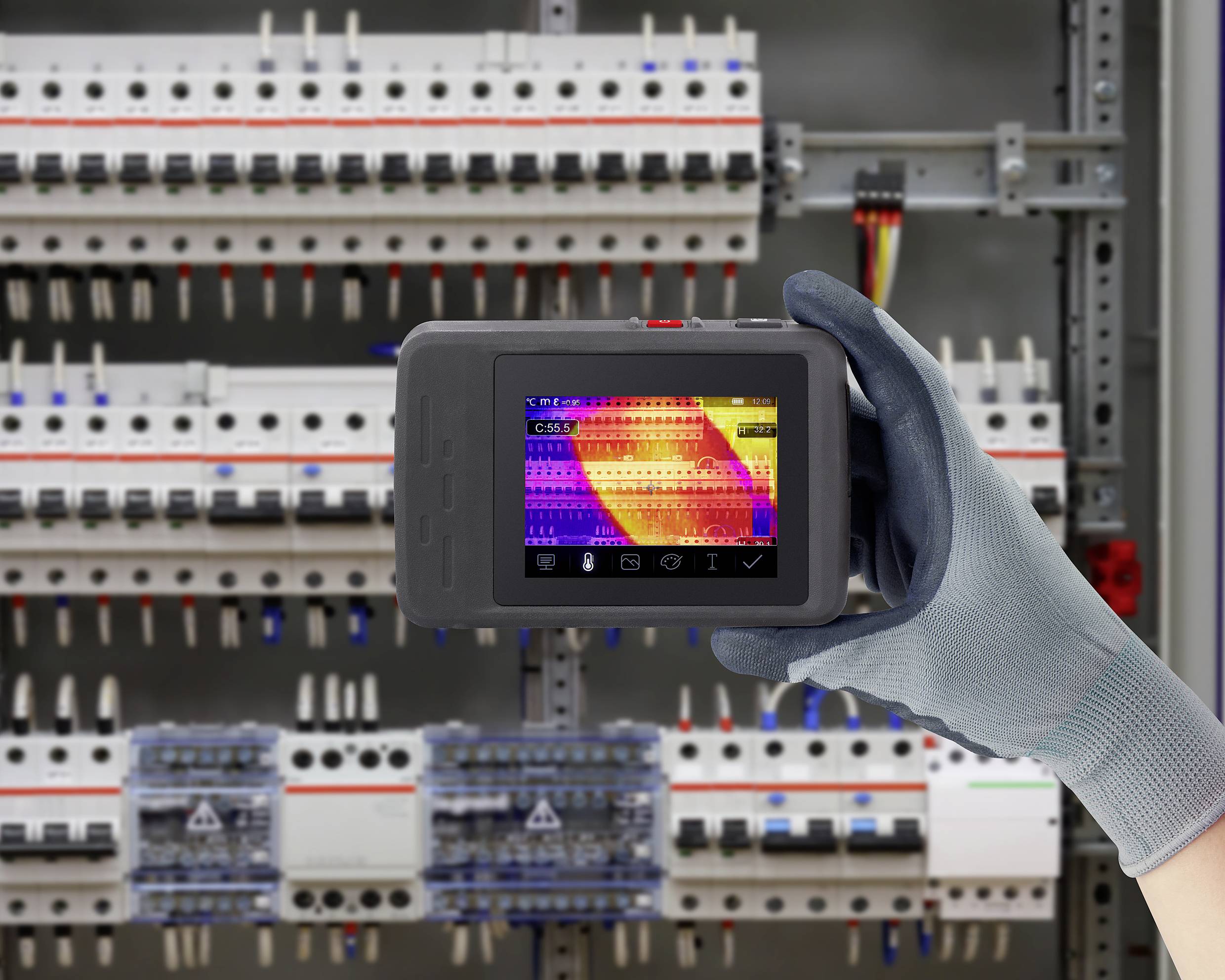 A person is holding a thermal imaging device in front of an electrical control panel. The display shows temperature variations in the electrical lines.