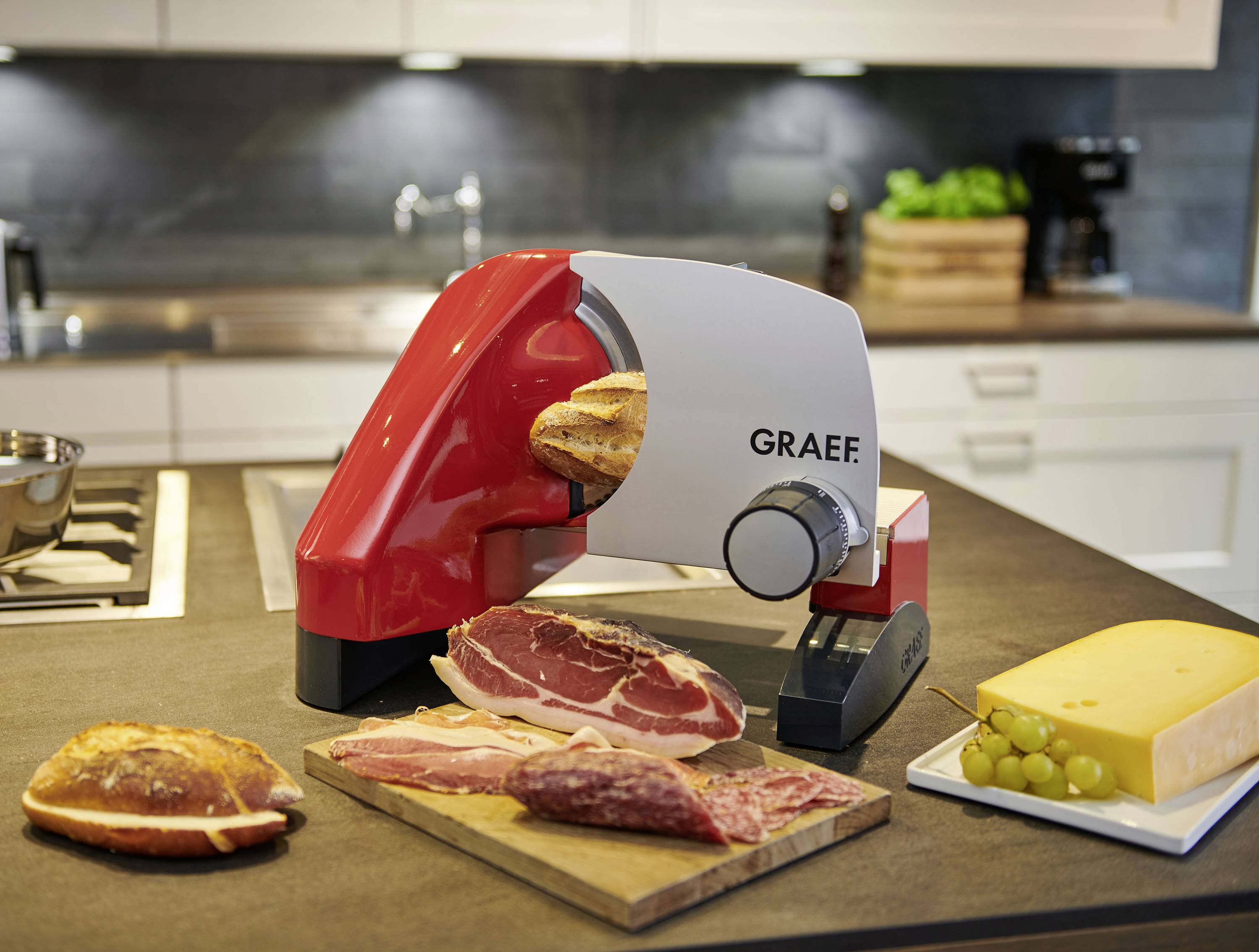 A red cold meat slicer on a kitchen worktop is cutting bread. Sausage and cheese are laid out in front of it. Kitchen appliances are visible in the background.