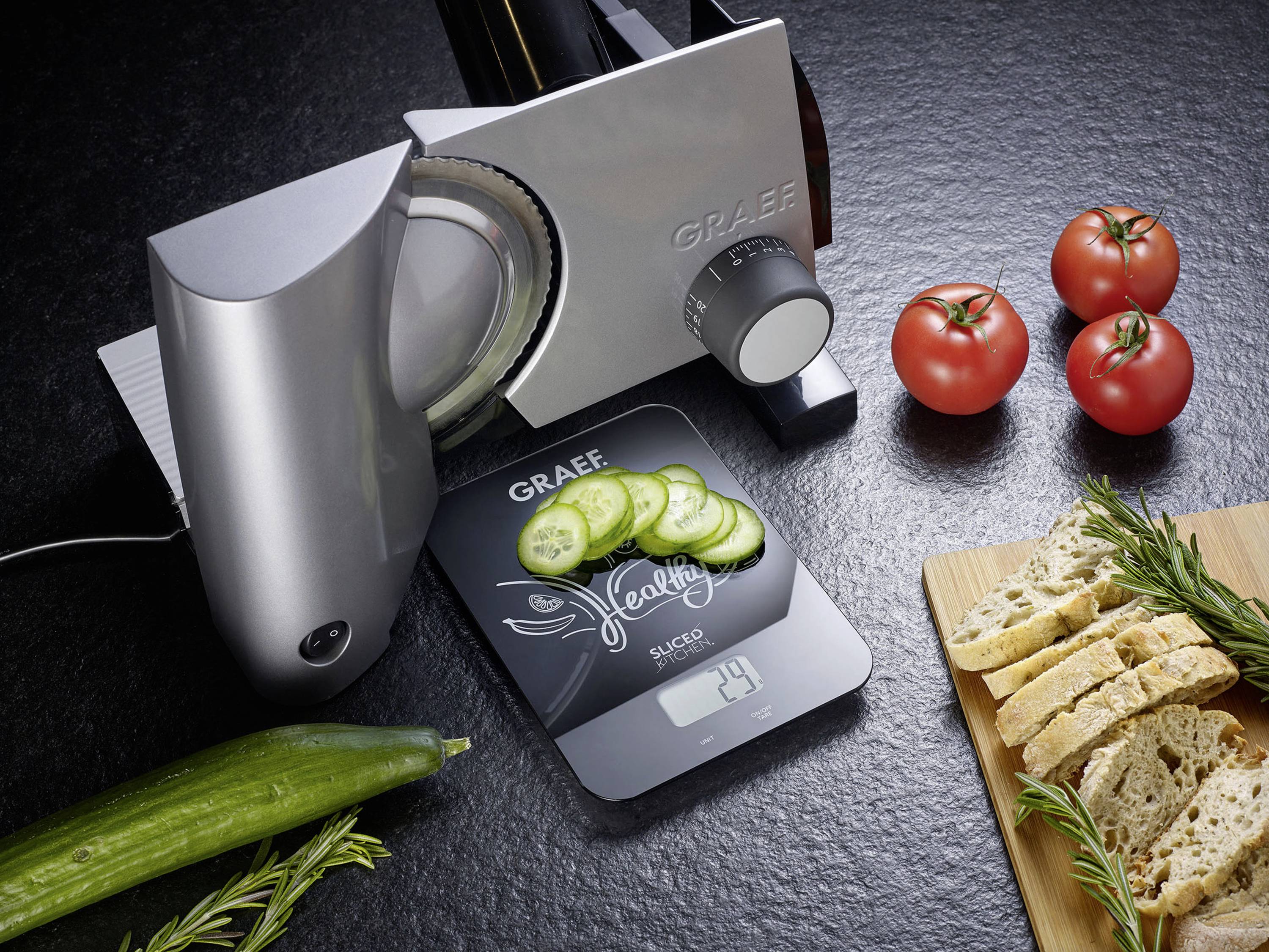 A silver universal slicer from Graef cuts cucumber slices resting on a digital scale. Beside it are tomatoes, bread and herbs on a worktop.