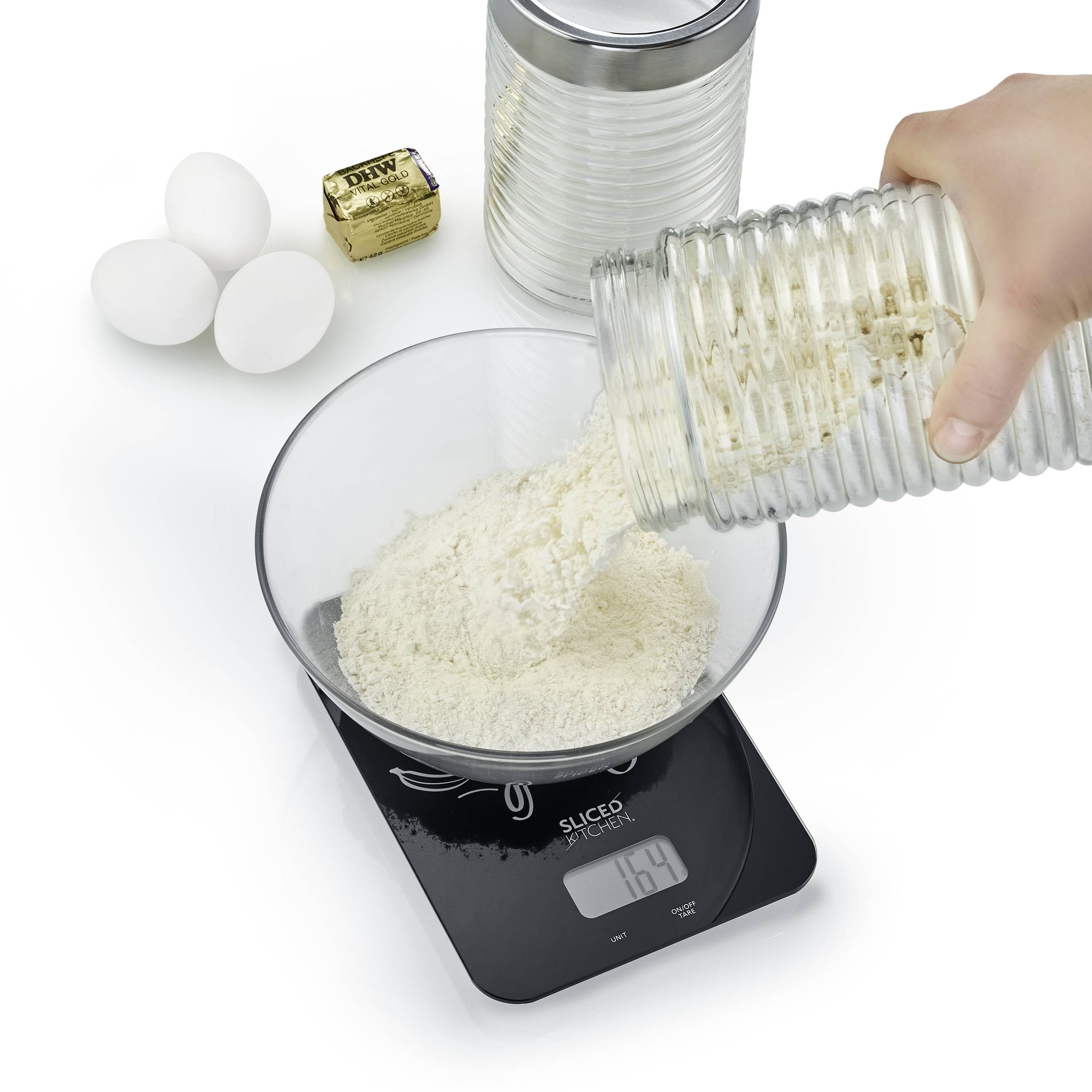A hand pours flour from a glass into a bowl on a digital scale. Three eggs and butter in gold foil are positioned beside it.