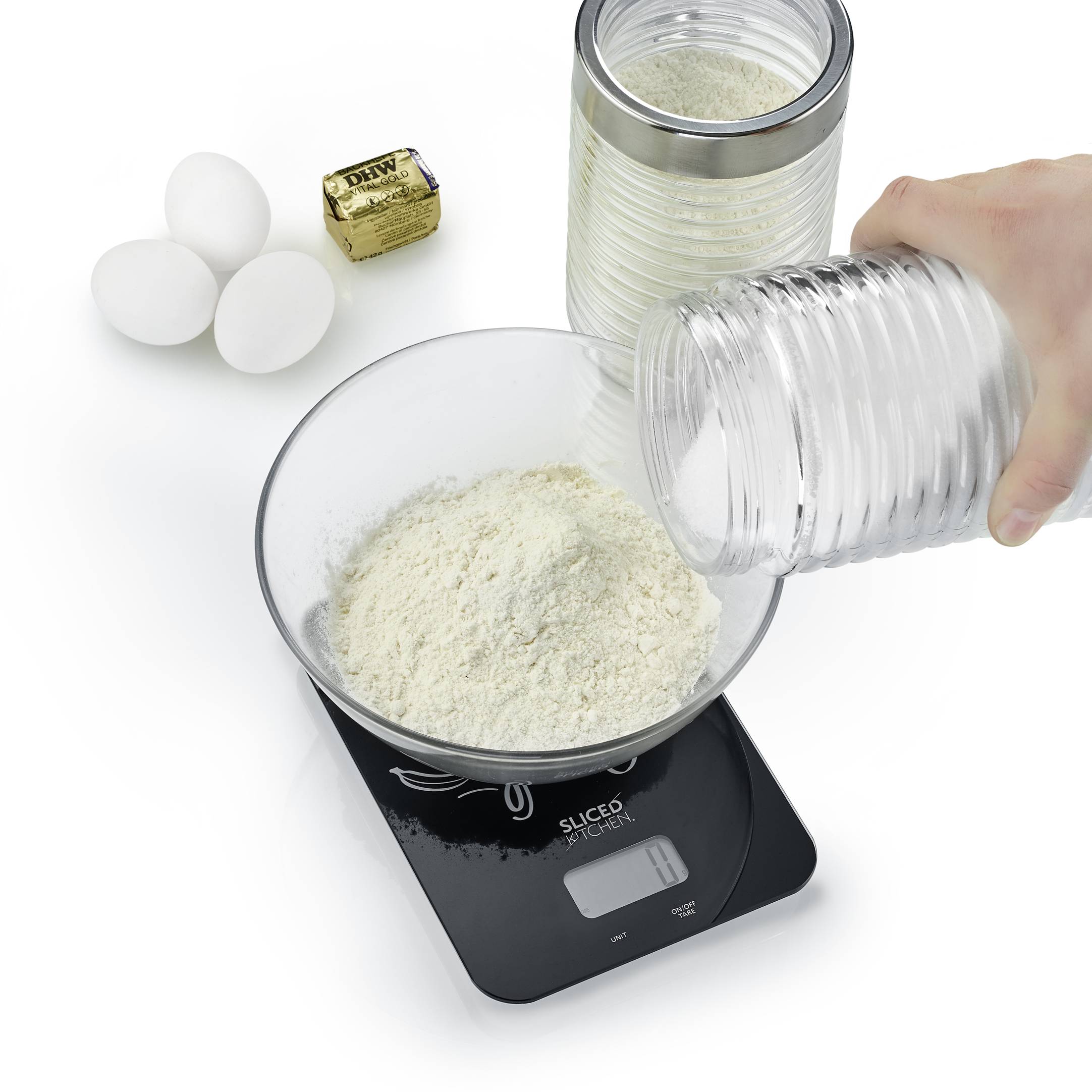 Hand pouring flour into a bowl on a digital scale. Beside it are three eggs, yeast, and a tin of flour.