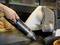 A hand holds a small vacuum cleaner, sucking up crumbs around a bread slicer. Bread rolls are visible in the background.