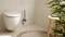 A modern bathroom with a white wall-mounted toilet, a toilet brush beside it, a wooden stool, and a plant arrangement.