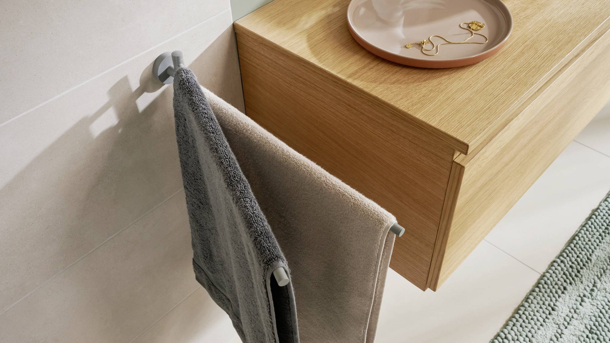 The image depicts a modern bathroom with a wall-mounted towel rail holding two towels, and a wooden shelf with a dish placed on it.