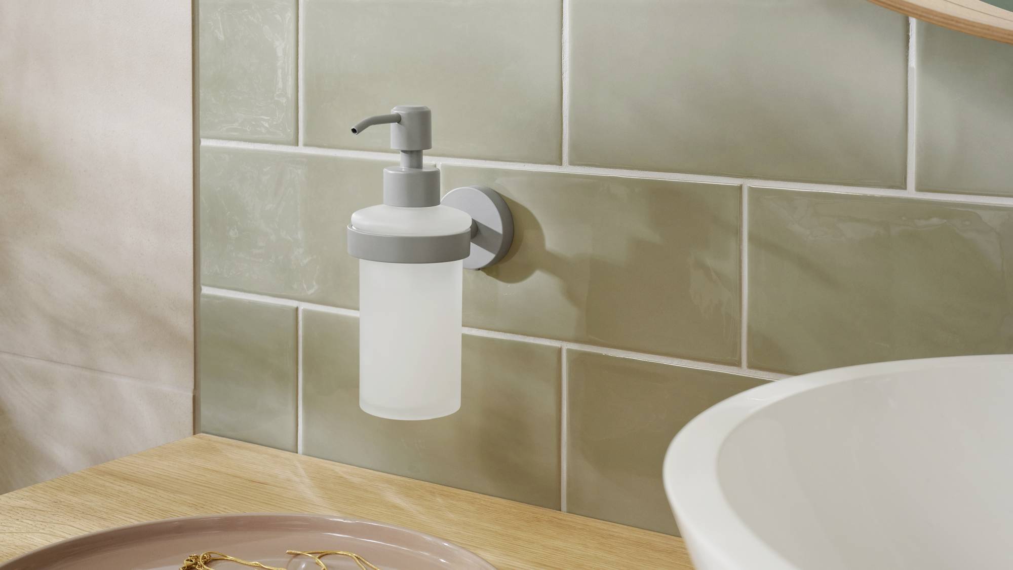 Wall-mounted soap dispenser made of frosted glass and metal on a green tiled wall, beside a washbasin with a wooden countertop.