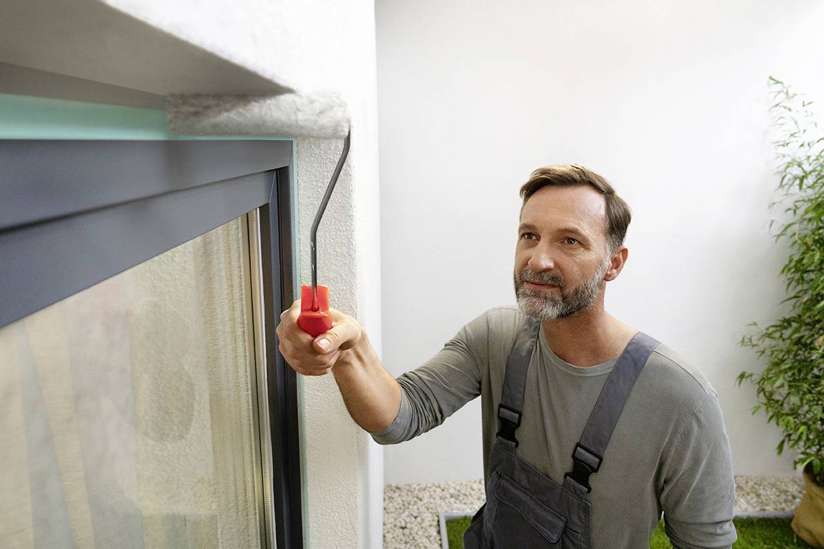 A man in workwear is painting an exterior wall white next to a window. Some greenery can be seen in the background.