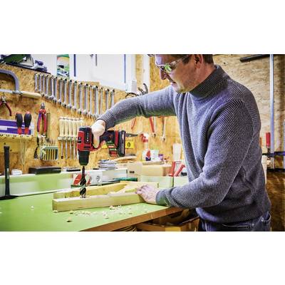 A man in a workshop is drilling into a piece of wood with a red drill, surrounded by tools on the wall.