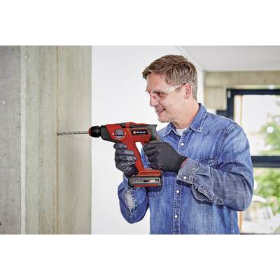 A man is drilling into a concrete wall with a red drill. He is wearing safety glasses and gloves, dressed in jeans.