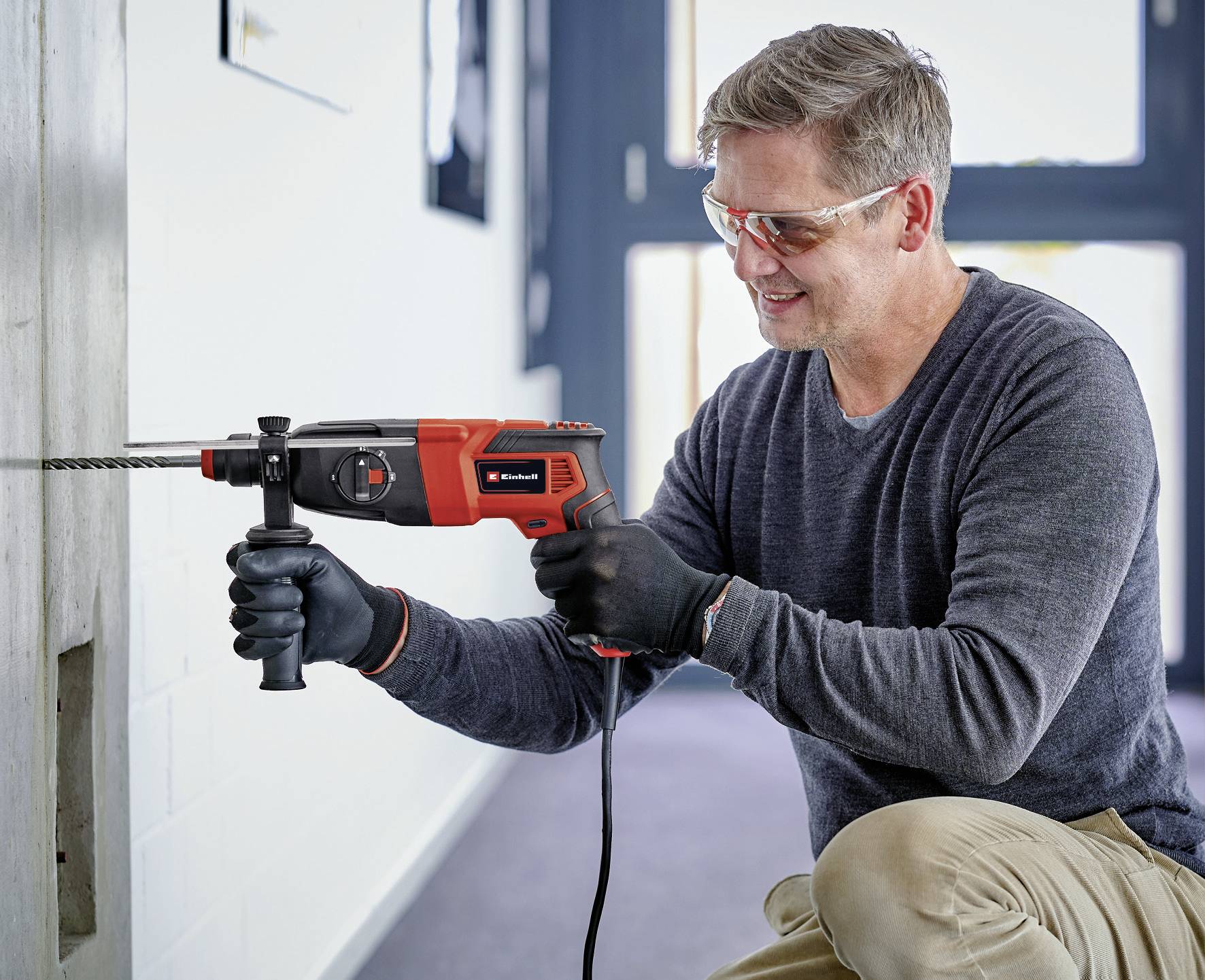 A man wearing safety glasses and gloves is drilling a hole in a concrete wall using an electric drill.