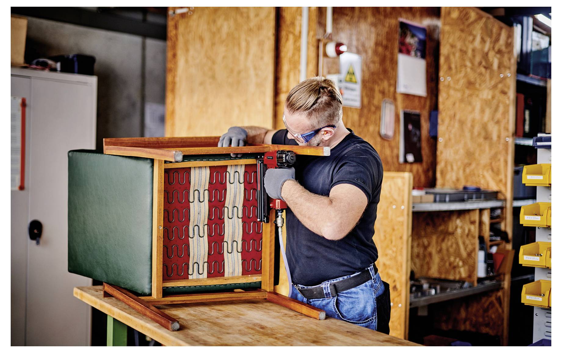 Person wearing safety goggles and gloves assembles a wooden cabinet or box on a workbench in a workshop.
