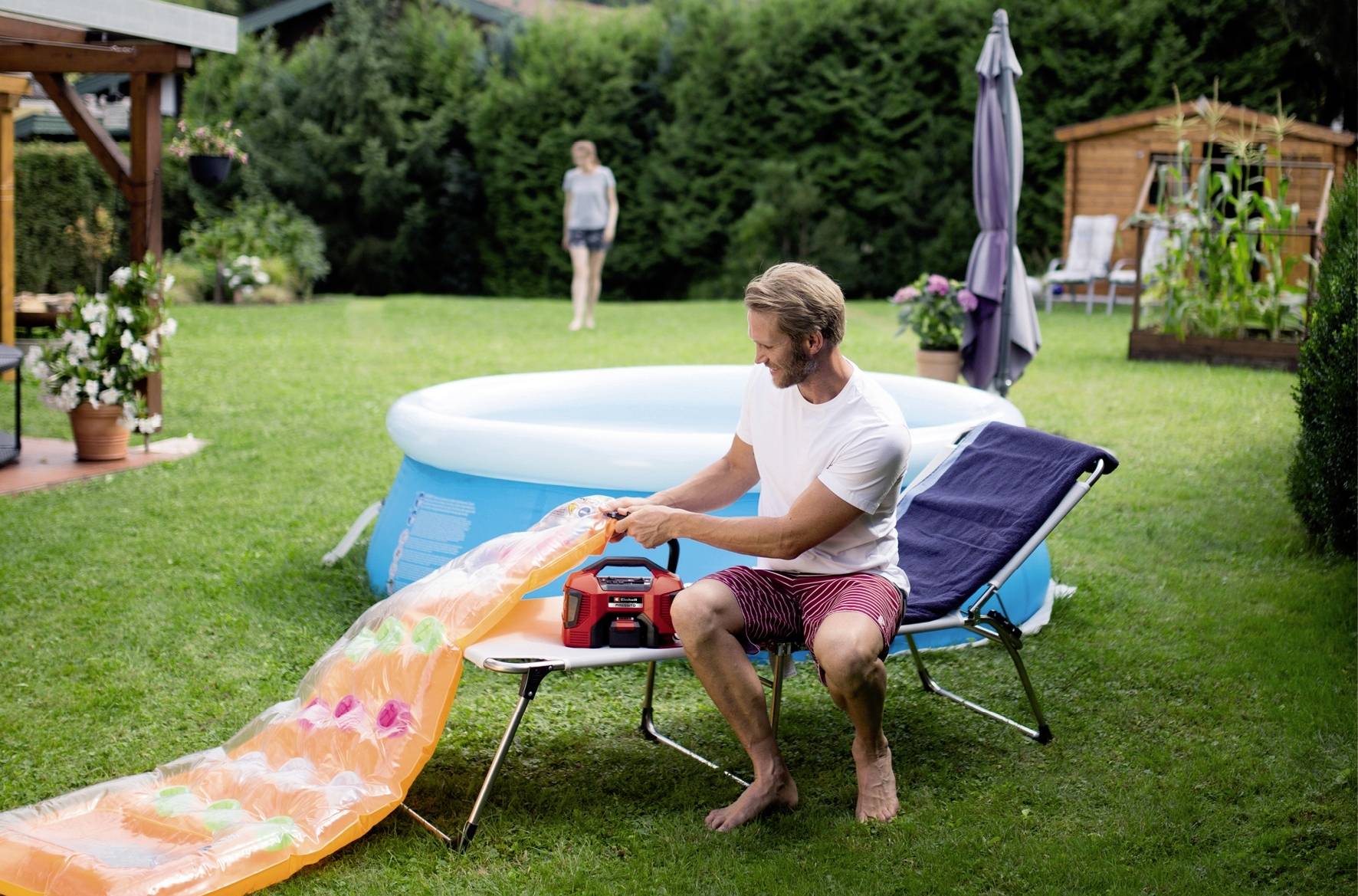 A man is inflating a colourful air mattress next to an inflatable pool in a garden. In the background, a woman is standing near a shed.