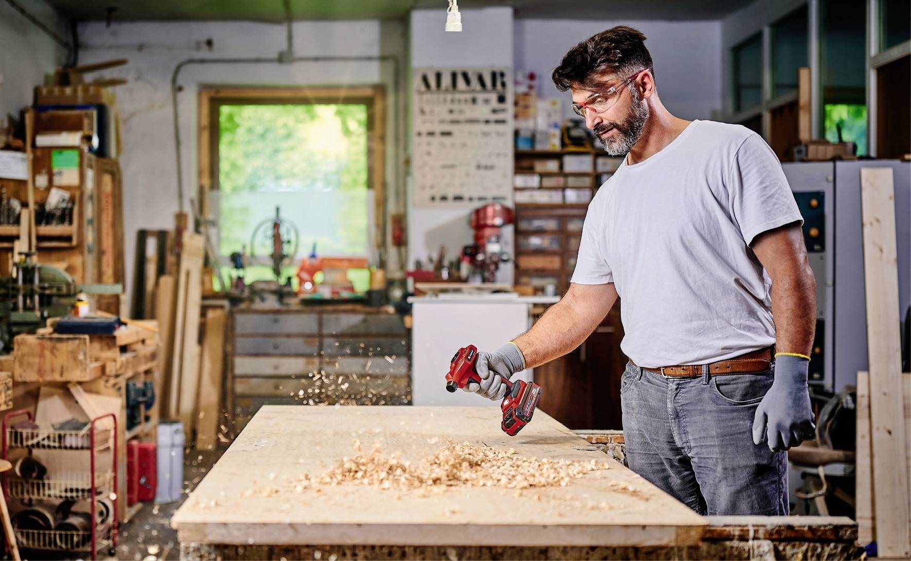 A man in a workshop is working with wood using a red and black machine, sawdust flying. Shelves and windows are visible in the background.