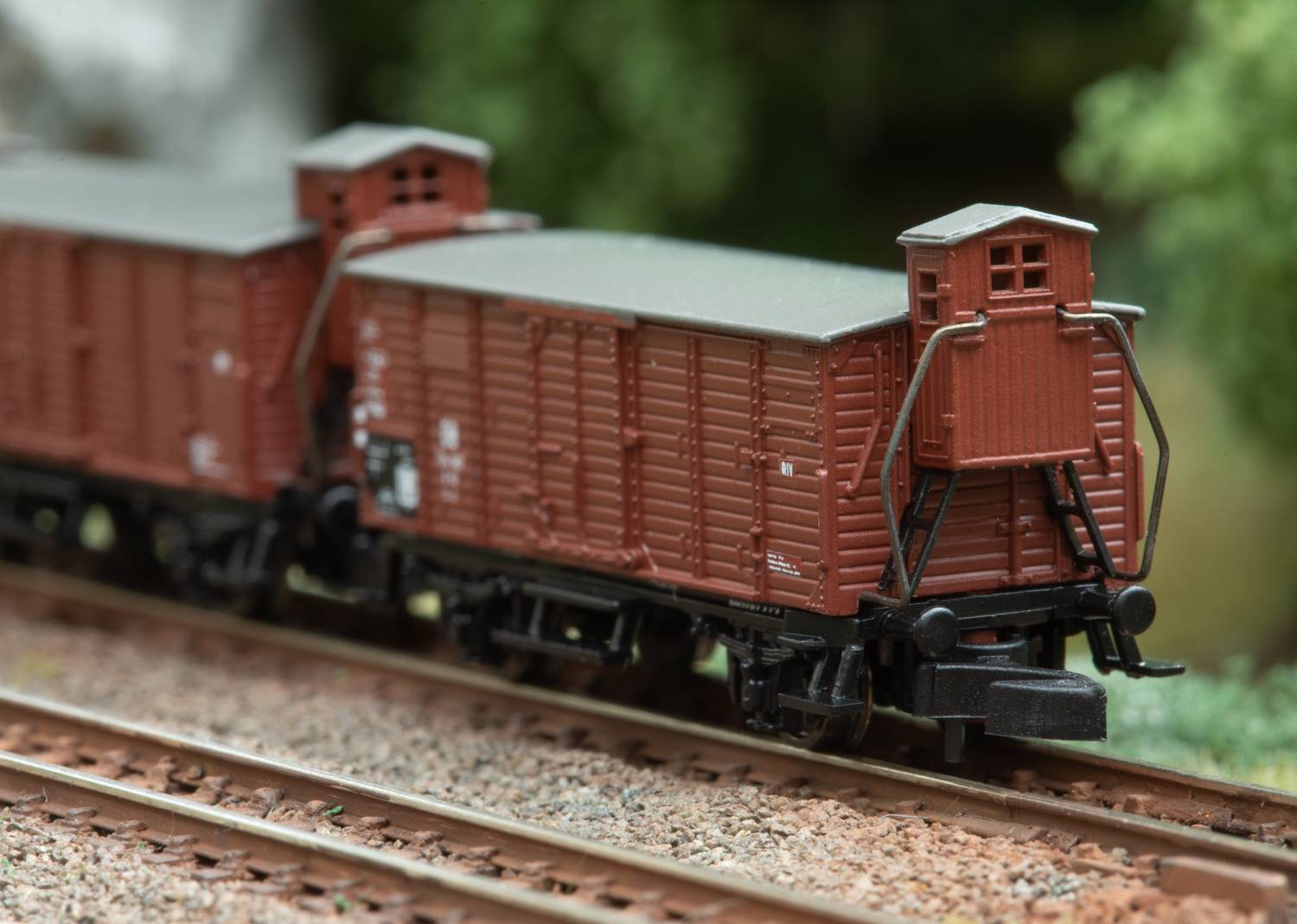 Two red freight wagons on tracks in a model railway setting, surrounded by a blurry green background, likely trees.