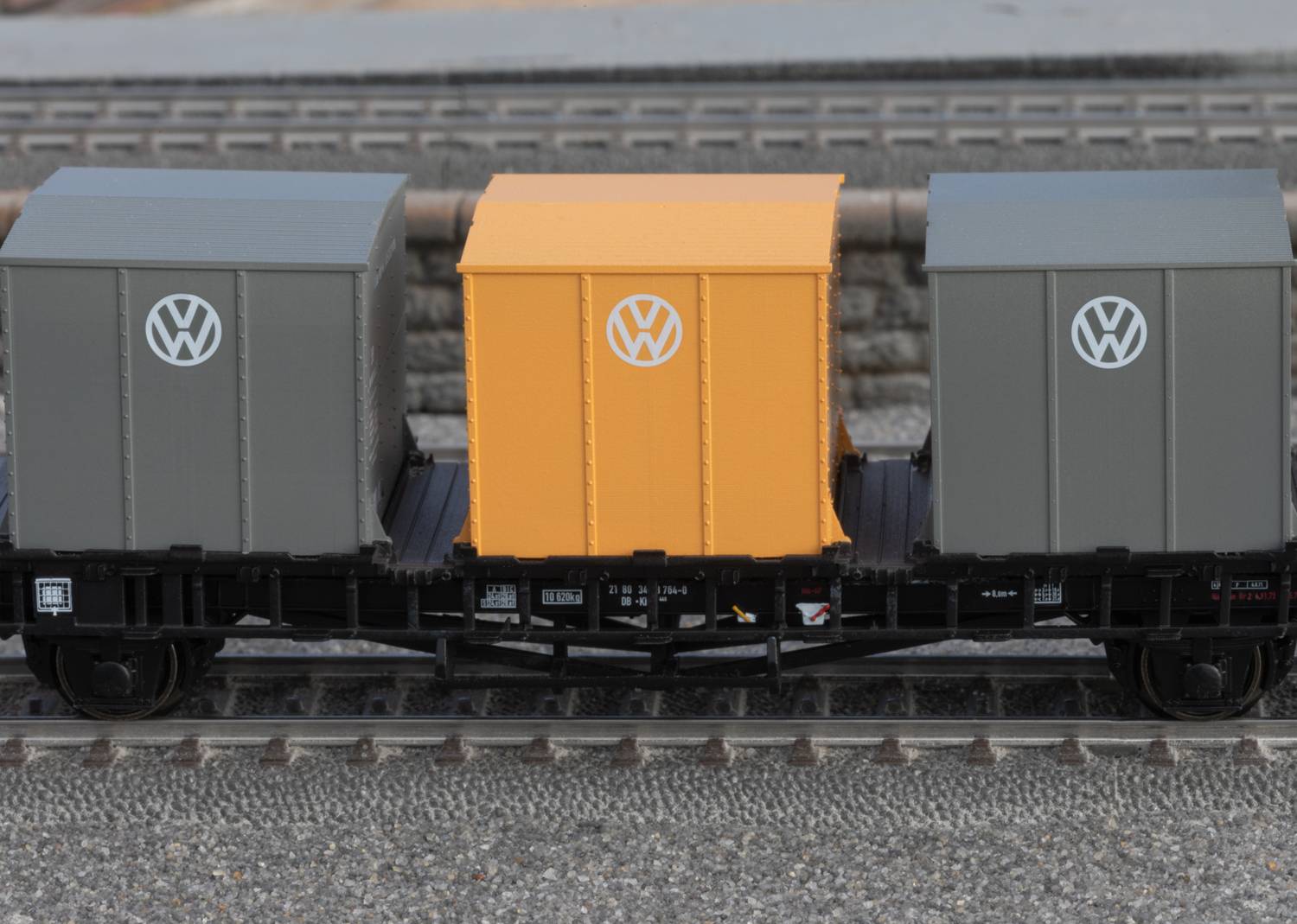 A model train with three freight containers, two grey ones with VW logos and a yellow one in the middle, is travelling along a model railway track.