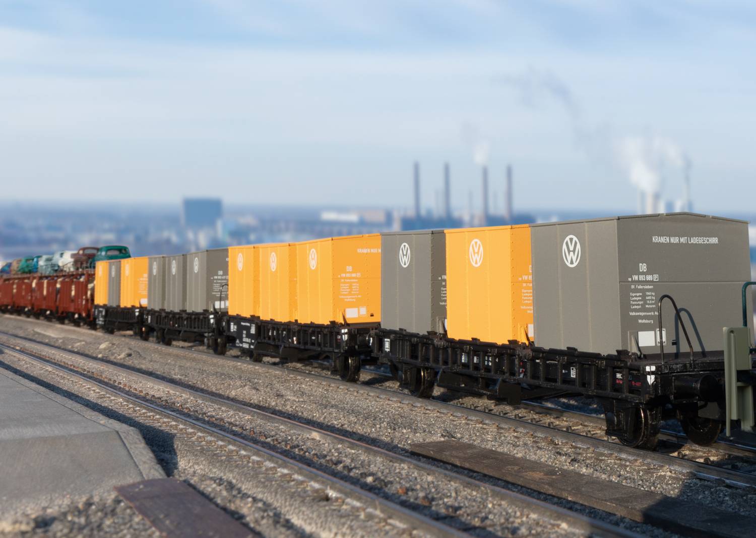 A railway line with several freight carriages, transporting containers with the VW logo. A factory can be seen in the background.